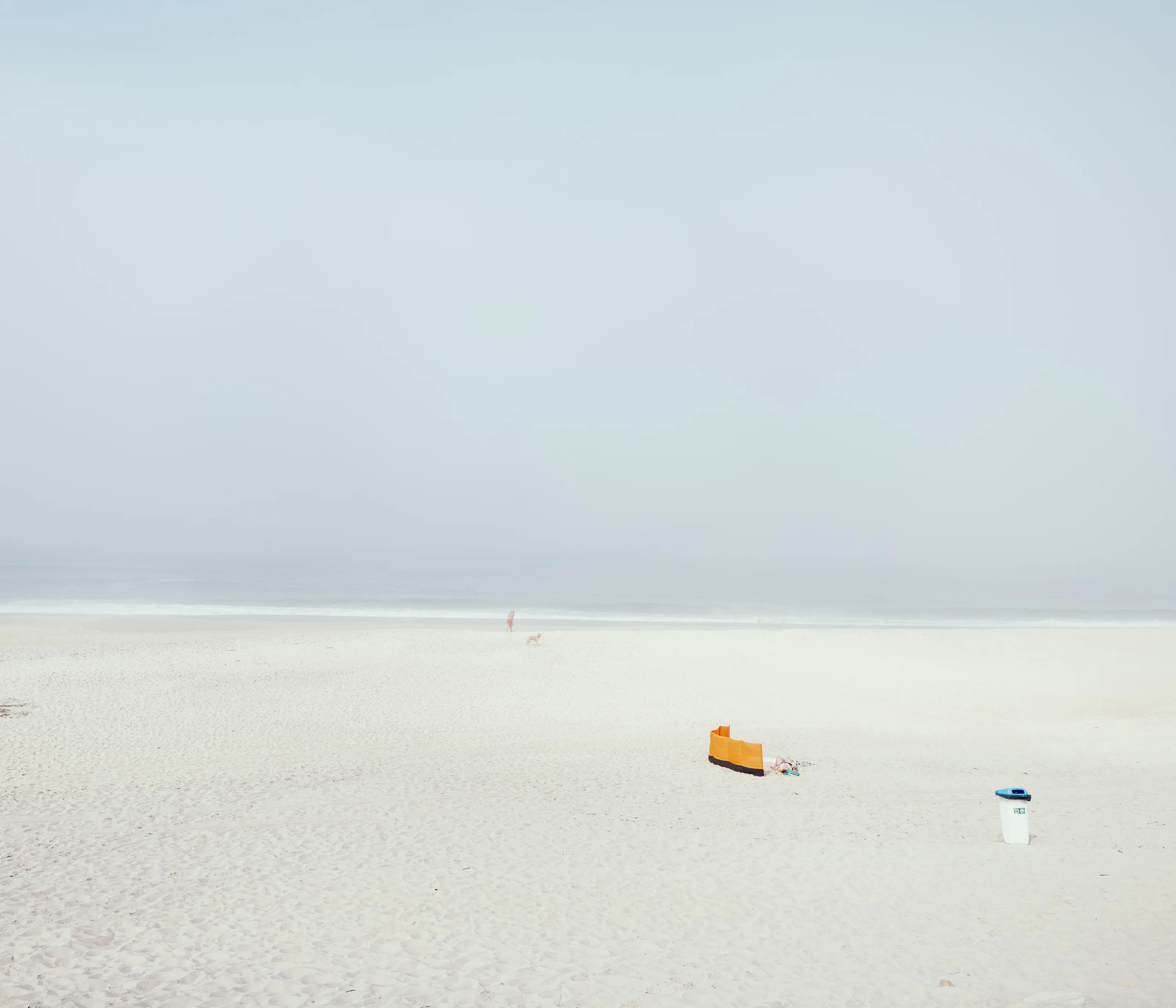 A minimalistic beach scene with a distant person and dog near the shoreline, and an orange windbreaker with a trash bin in the foreground.