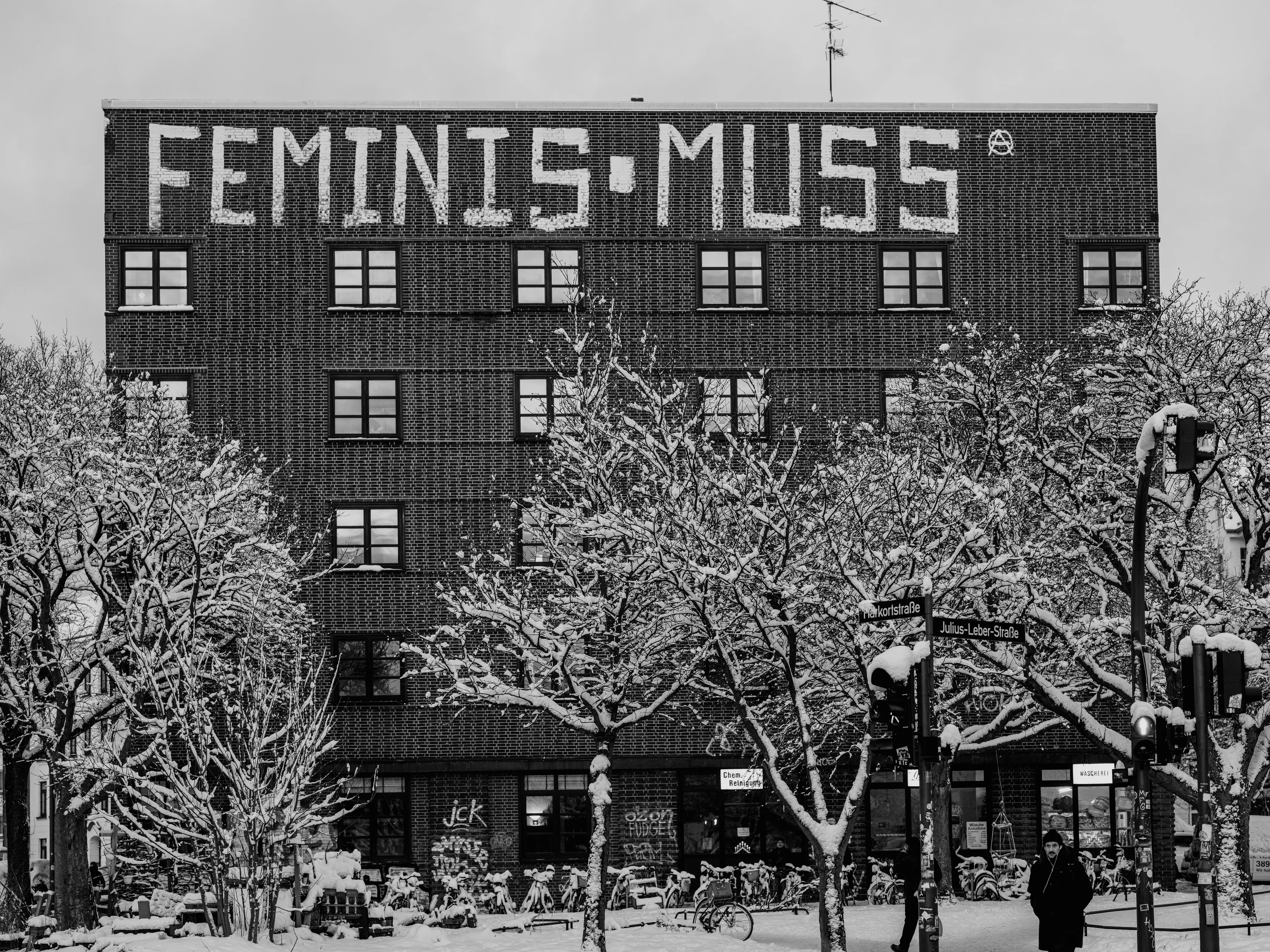 A brick building with large text 'FEMINISMUSS' on the facade, surrounded by snow-covered trees.