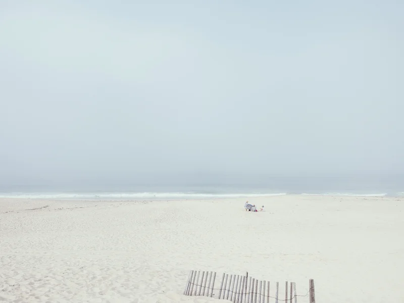 A sparse beach scene with a thin, uneven wooden fence in the foreground and a small group of people sitting near the water's edge.