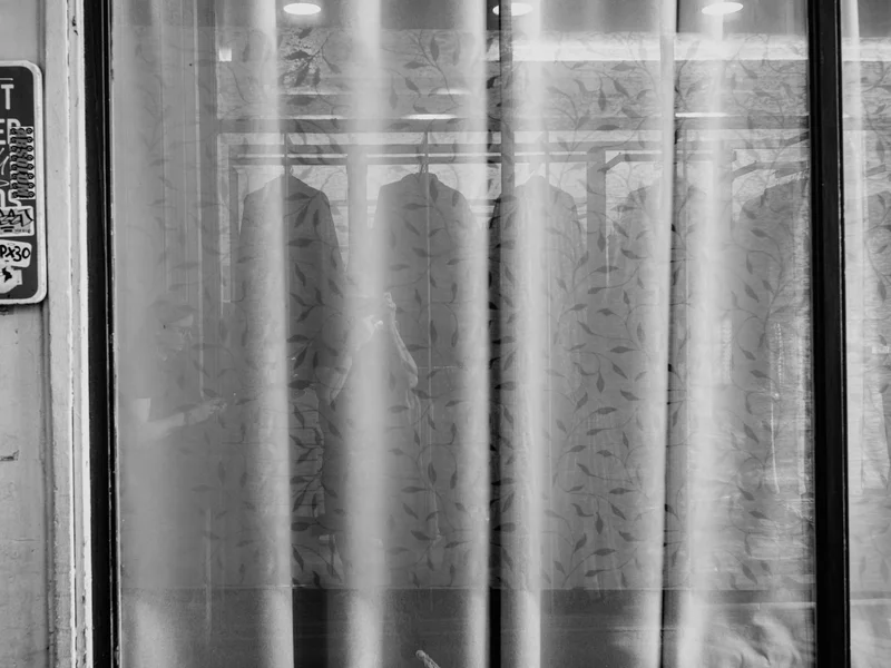 Black and white photo of a patterned curtain with shadows of hanging clothes behind, and partially obscured urban signs on the left.