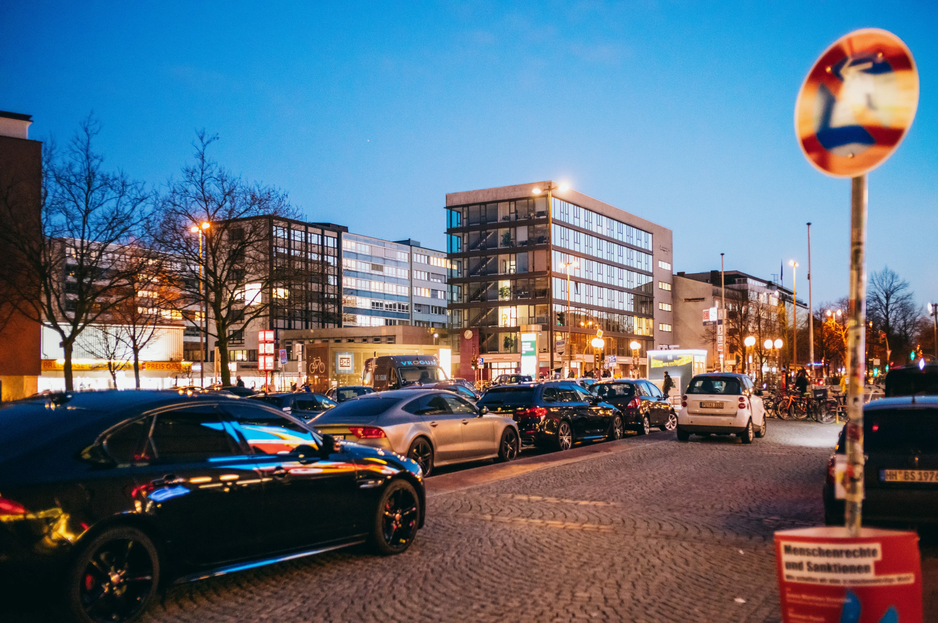 City street at dusk with parked cars and modern buildings in the background.