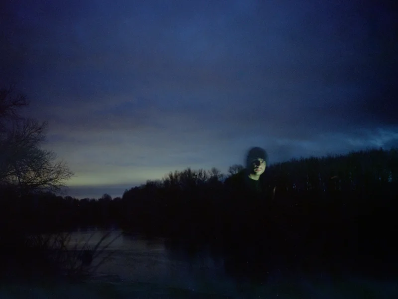 A person with a faintly illuminated face stands in front of a dark, twilight landscape with trees and water.