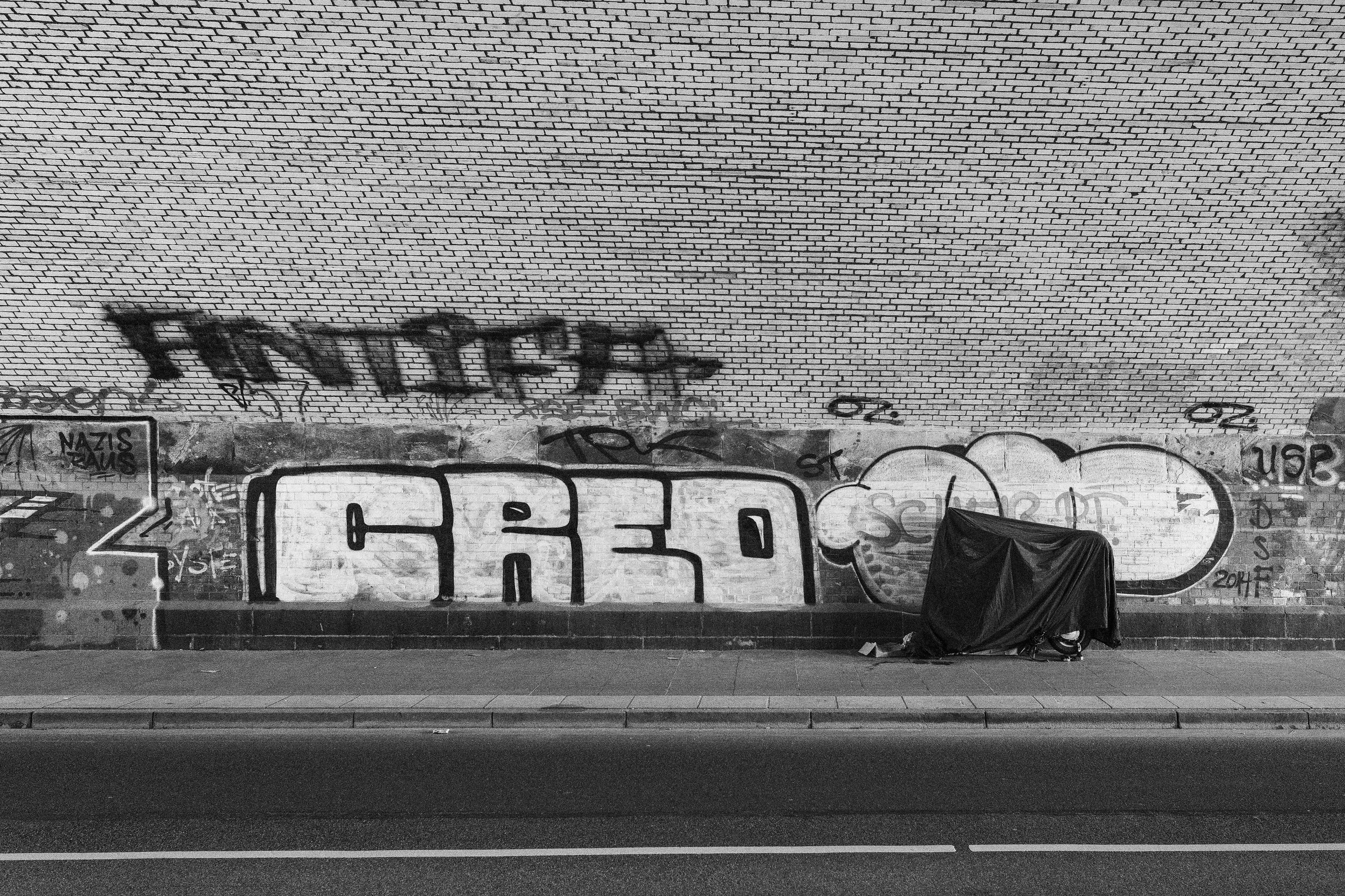 Black and white graffiti on a brick wall above a covered object on the sidewalk.
