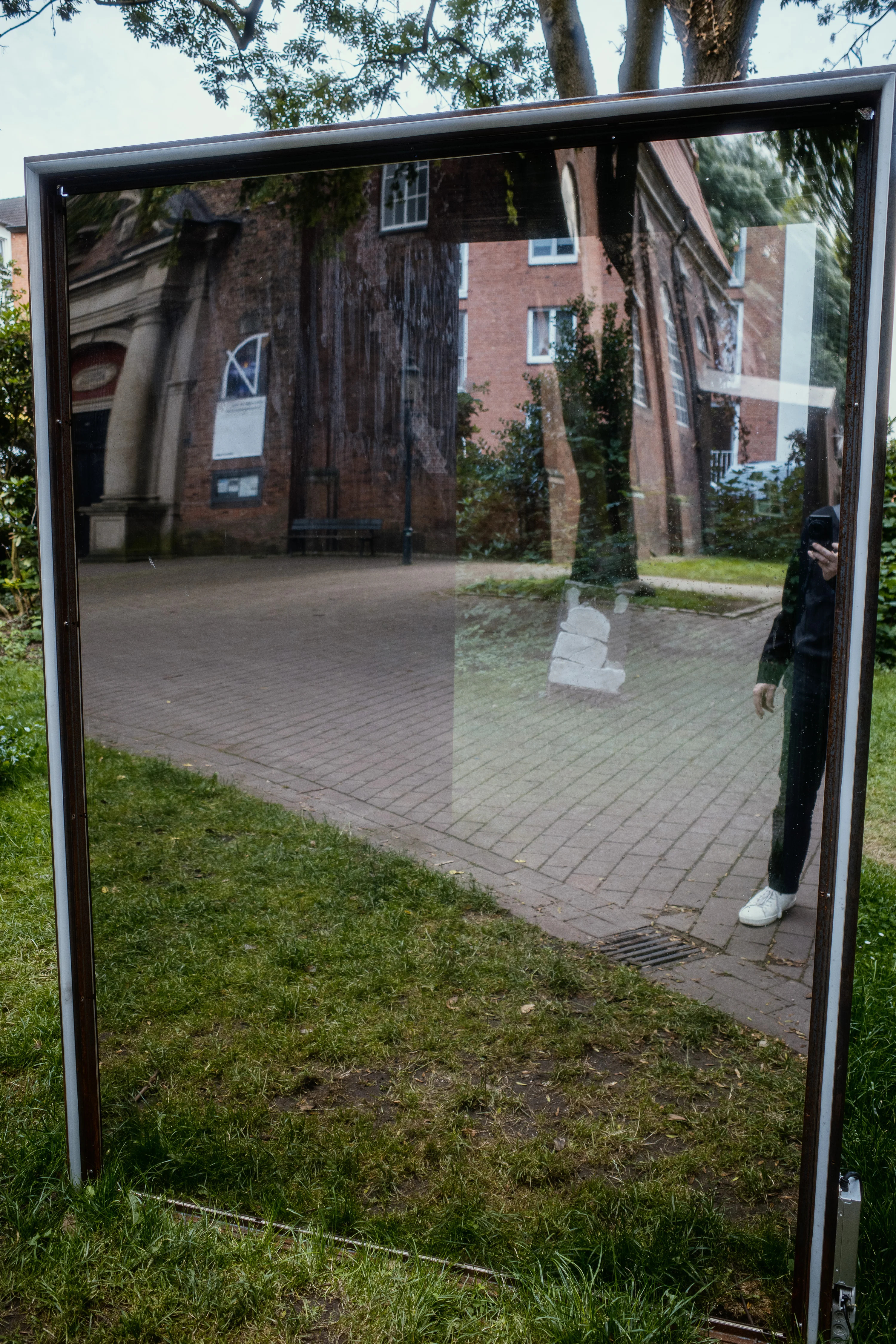 A reflective glass panel displaying a distorted reflection of a brick building and trees, with a person partially visible on the right.