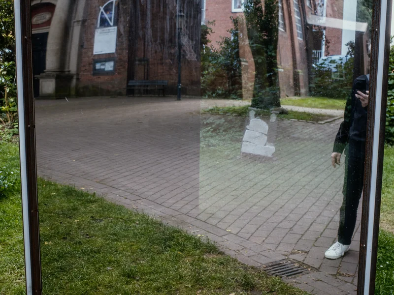 A reflective glass panel displaying a distorted reflection of a brick building and trees, with a person partially visible on the right.