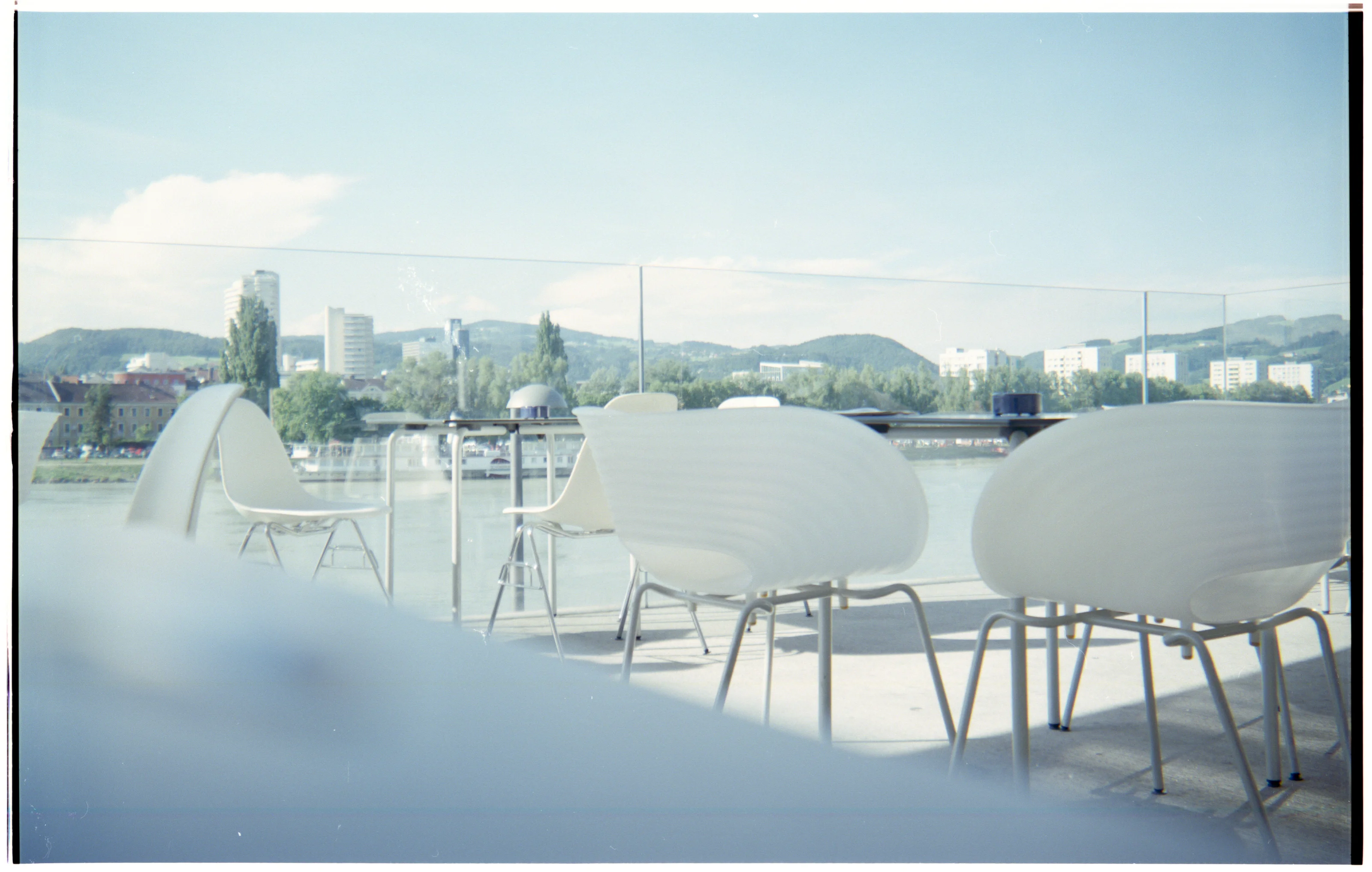 Outdoor seating area with white chairs and a glass railing overlooking a river and cityscape.
