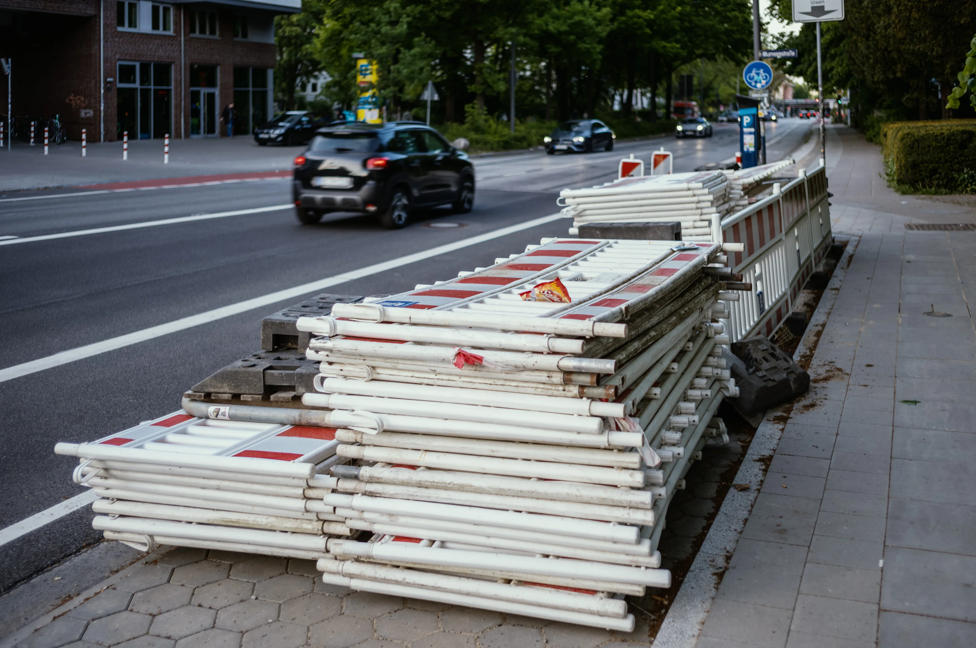 Stack of white and red construction barriers on a sidewalk beside a road with passing cars.
