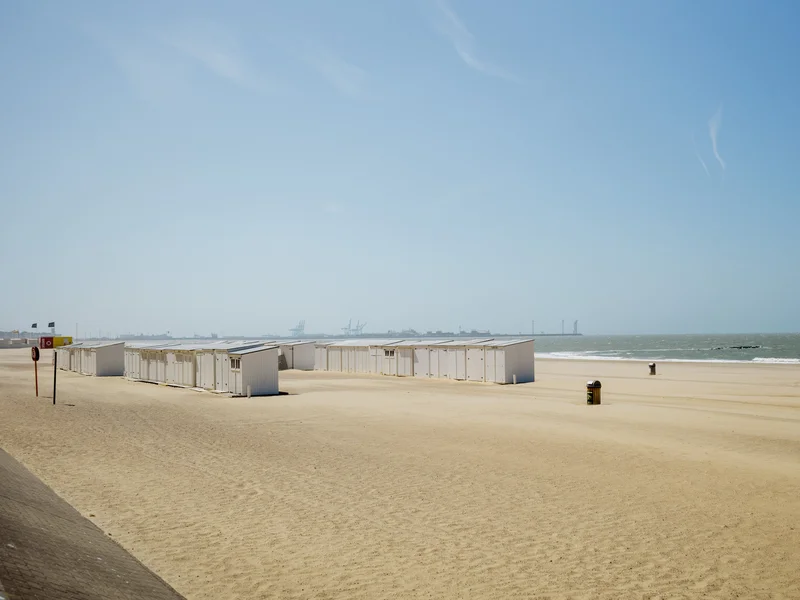 Sandy beach with row of white cabins near the shoreline under a clear blue sky.