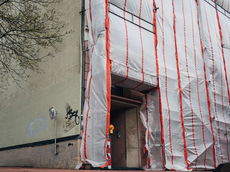 Building facade covered with white and red striped protective wrap next to a grafittied wall.