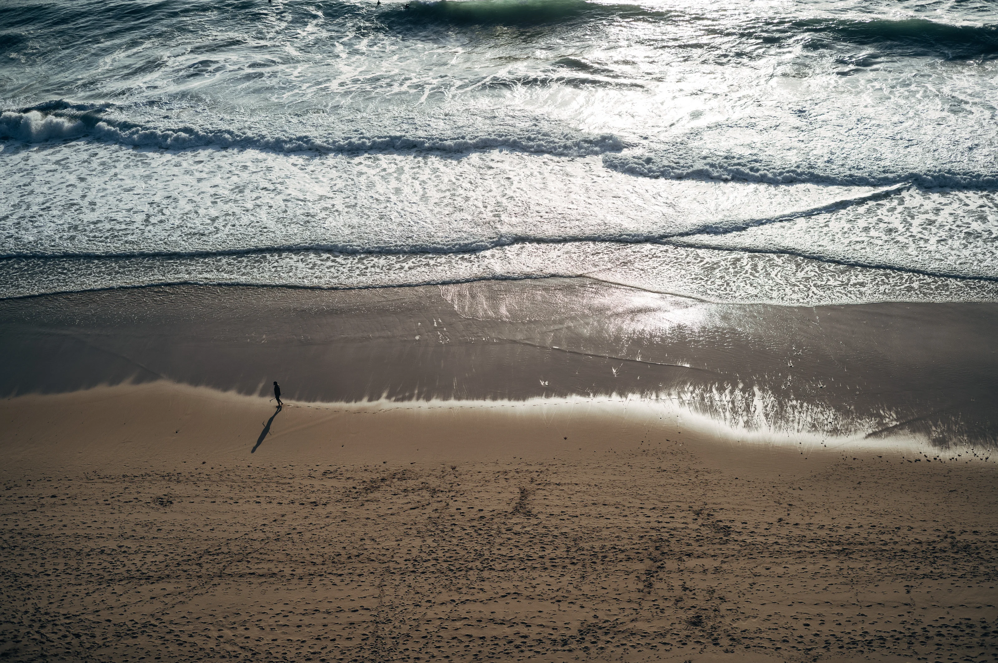 A lone person walks along a sandy beach with ocean waves rolling in.