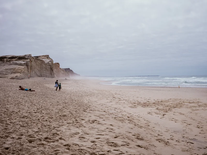 People walking along a wide, sandy beach with cliffs in the distance.