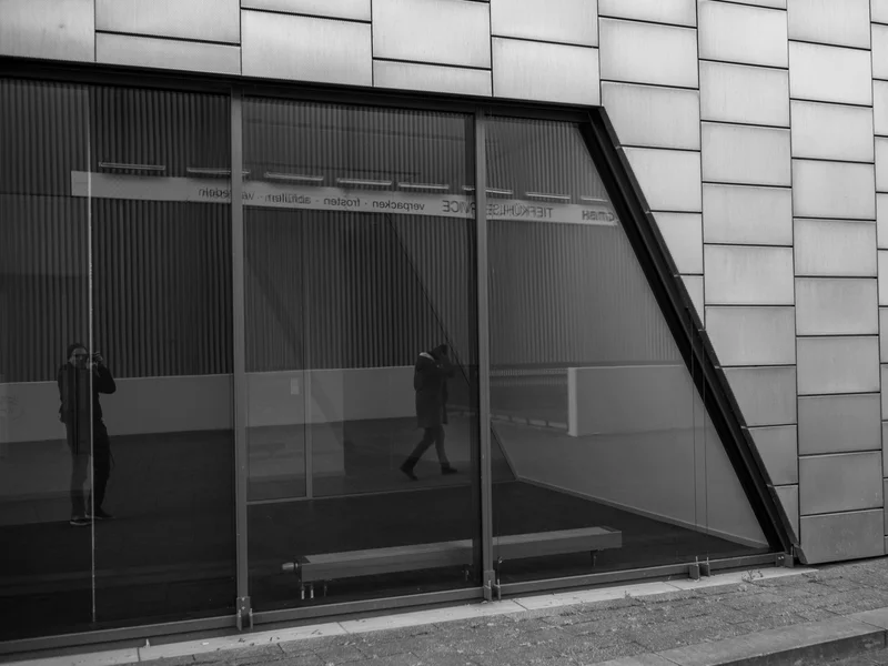 Black and white photo of a building facade with reflective windows and a person walking, reflected in the glass.