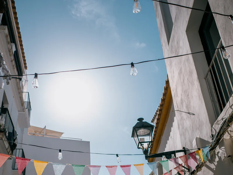 Narrow alleyway with colorful bunting and light bulbs strung between buildings under a clear blue sky.