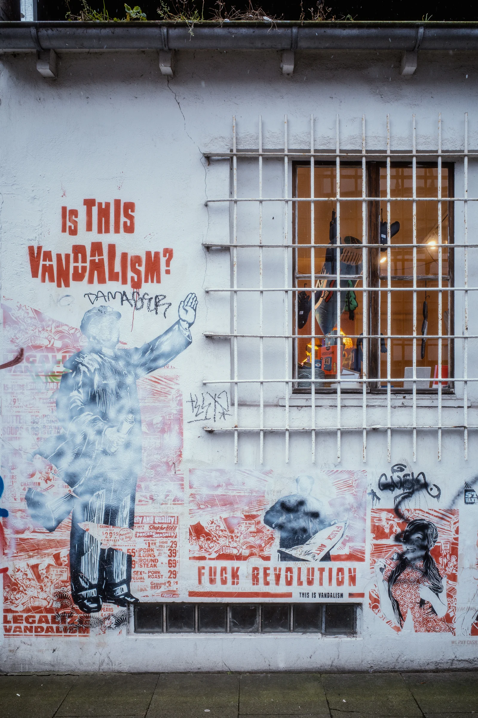 Street art on a white wall reading 'IS THIS VANDALISM?' above pasted posters, one captioned 'FUCK REVOLUTION — THIS IS VANDALISM', beside a barred window.