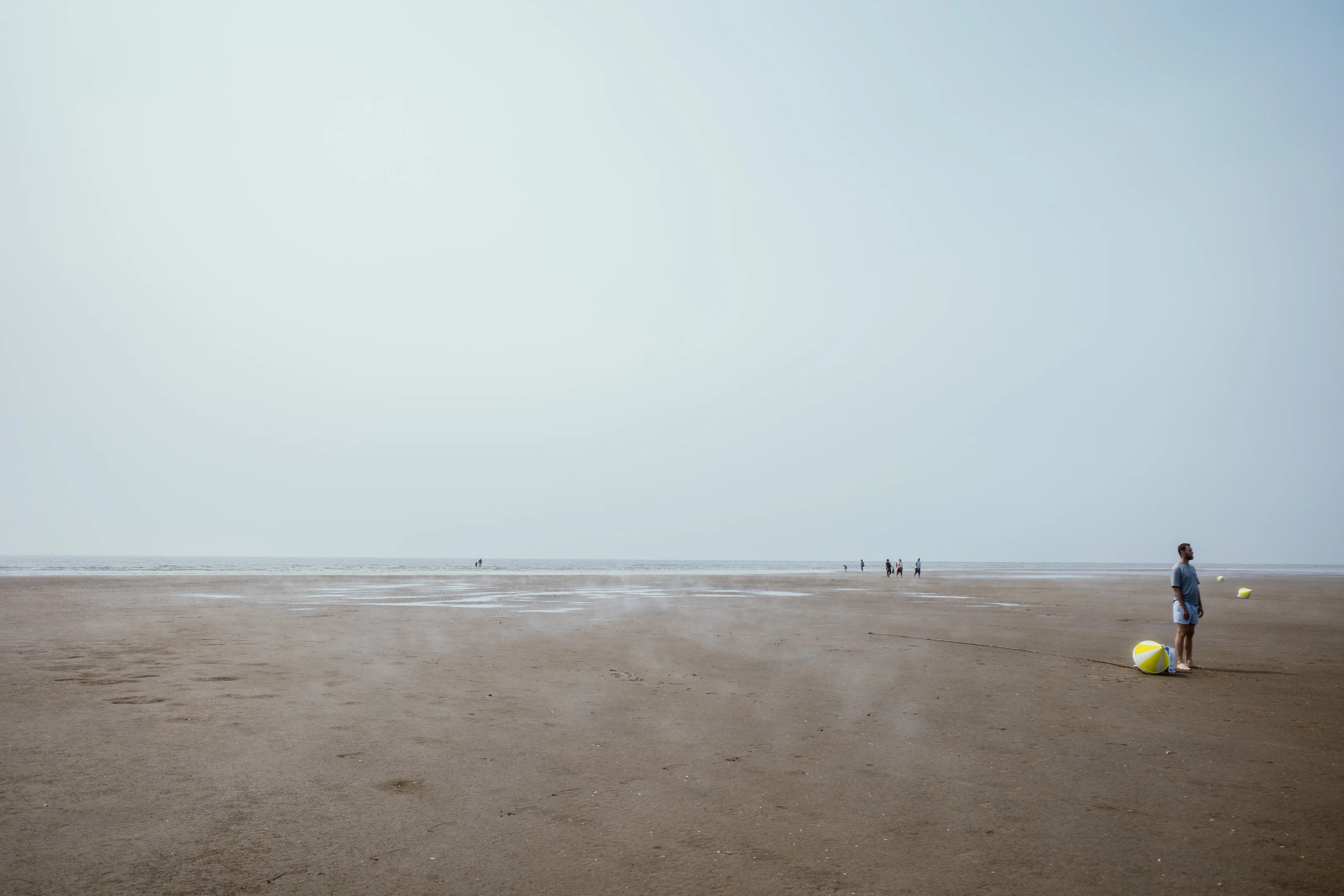 A person stands with a beach ball on a vast sandy beach with a hazy sky.