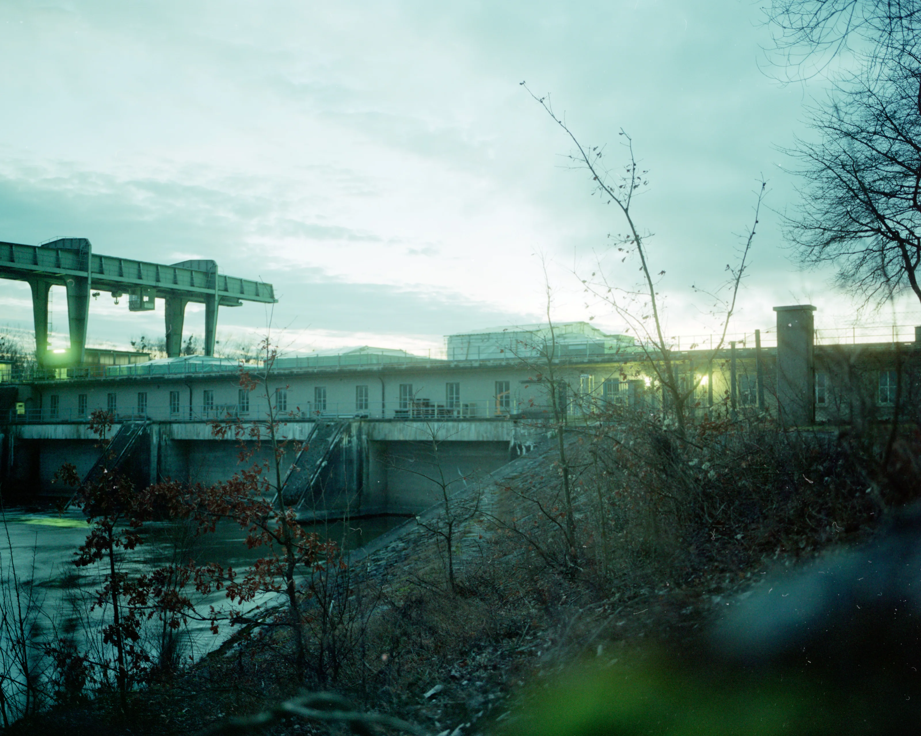 Industrial building and structures silhouetted against a cloudy sky with bare trees in the foreground.