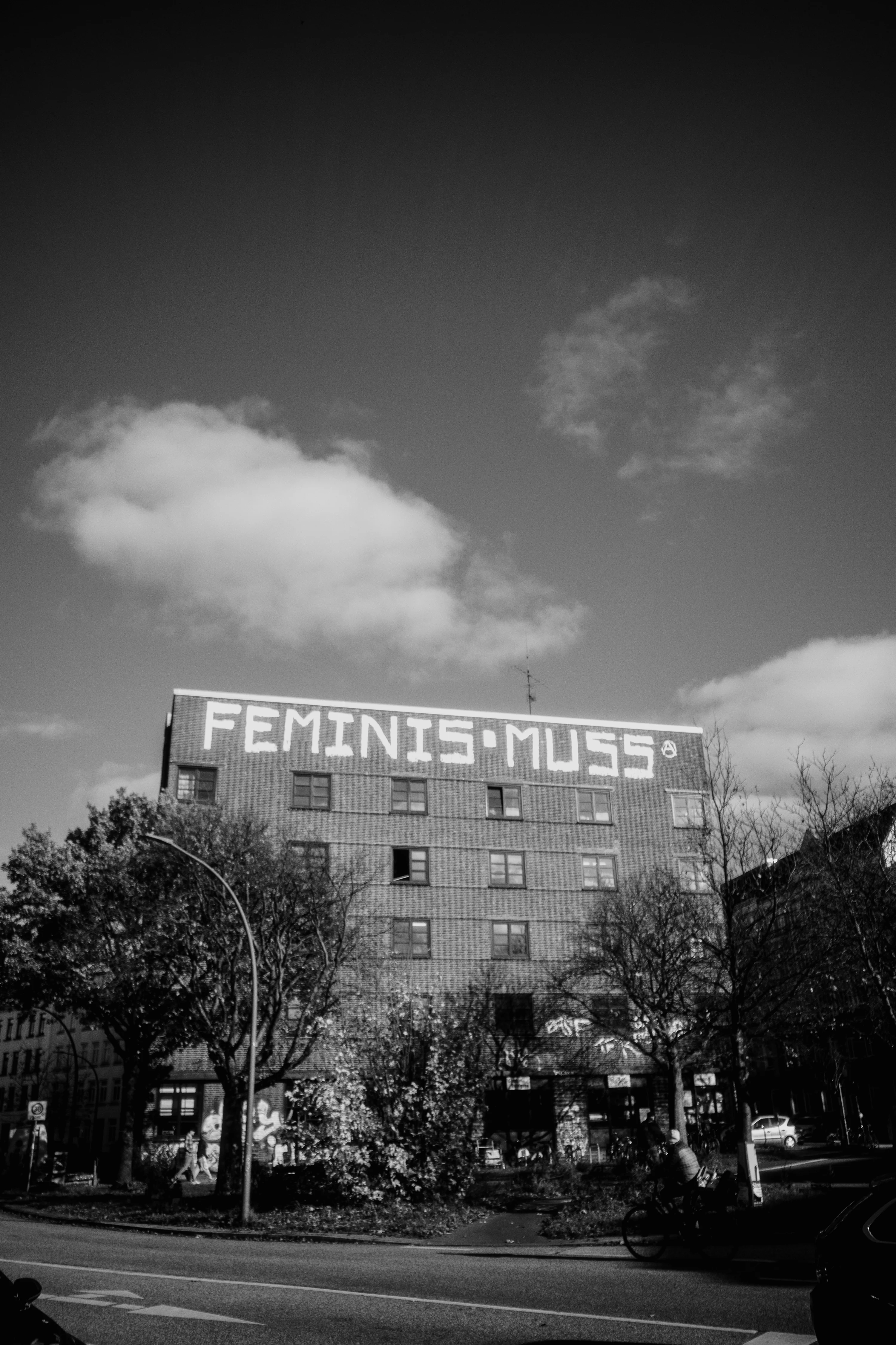 A building with graffiti reading 'FEMINISMUSS' on the top, under a cloudy sky.