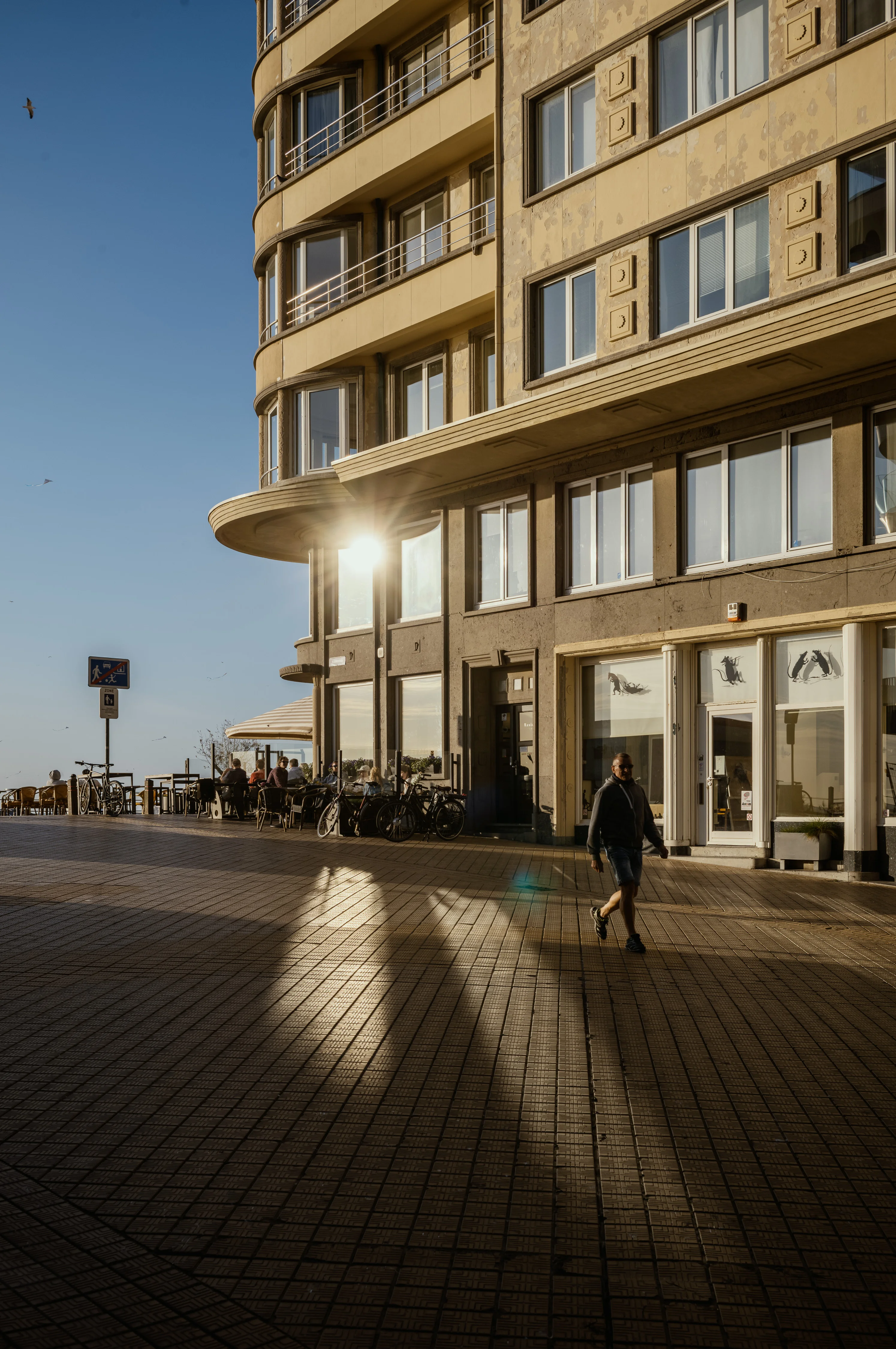 A person walks past a sunlit corner of a building with outdoor seating.