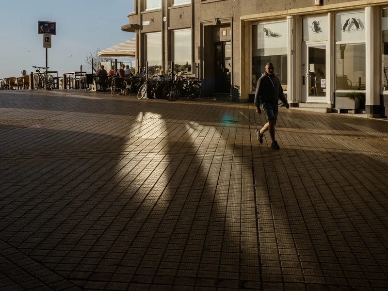 A person walks past a sunlit corner of a building with outdoor seating.