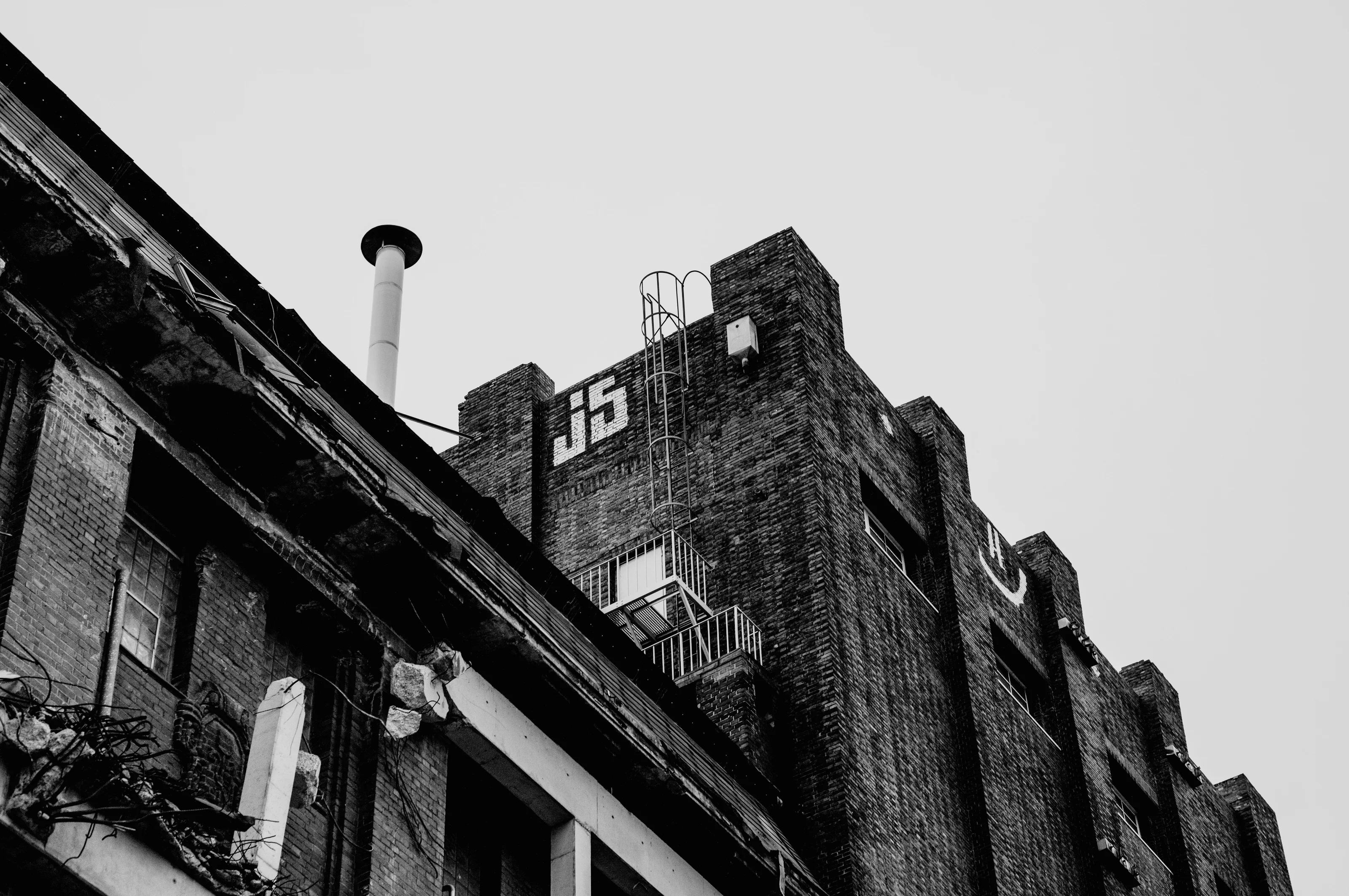 Black and white photo of an old industrial building with a chimney and 'J15' marked on the brick wall.
