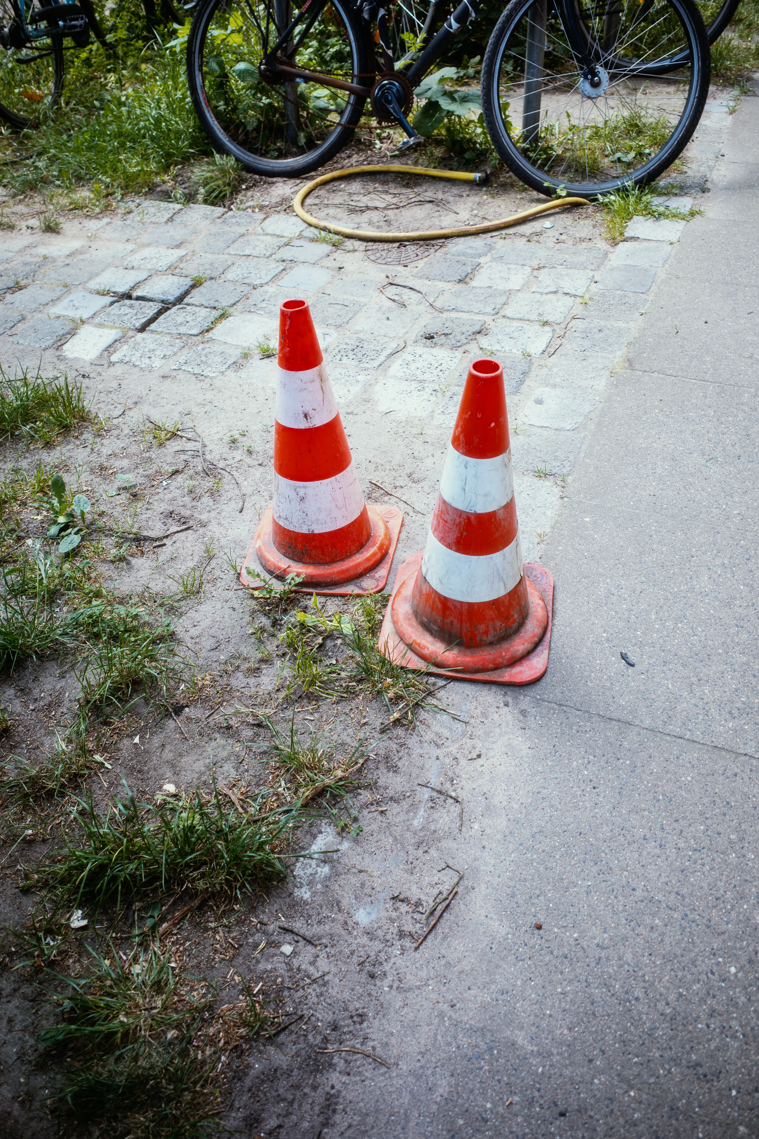 Two orange and white traffic cones on a patch of dirt and grass near a paved path and bicycle.