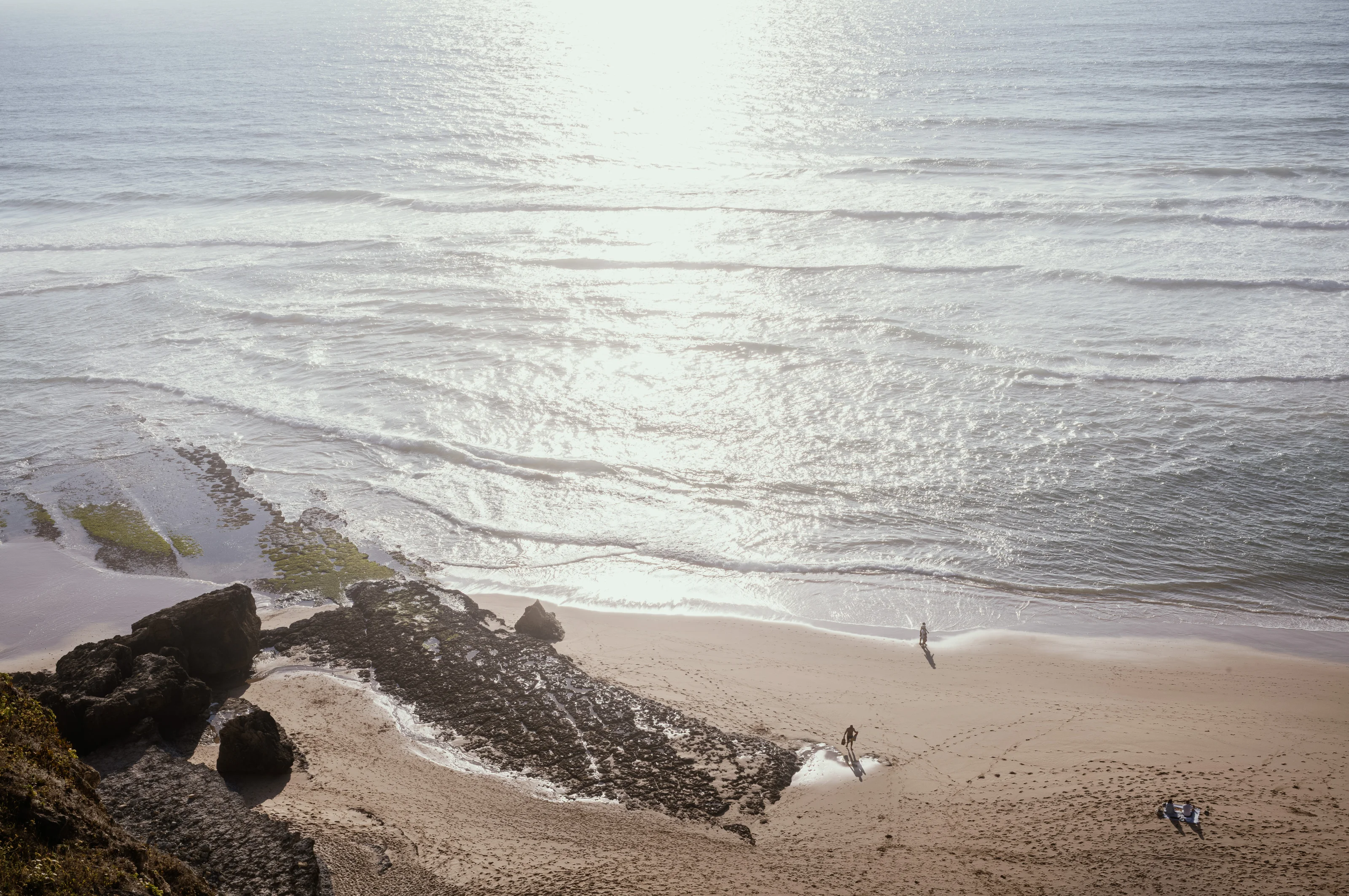 Beach with two people walking on the sand near the shore and a surfboard on the ground.