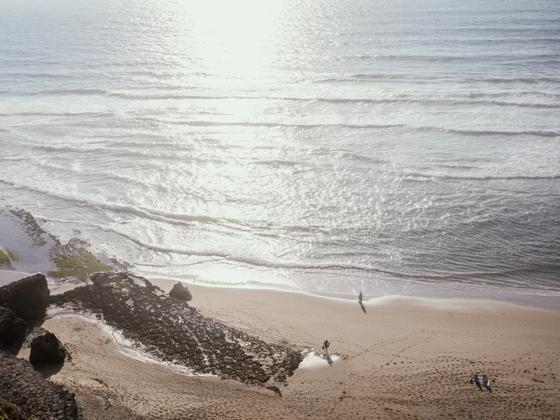 Beach with two people walking on the sand near the shore and a surfboard on the ground.