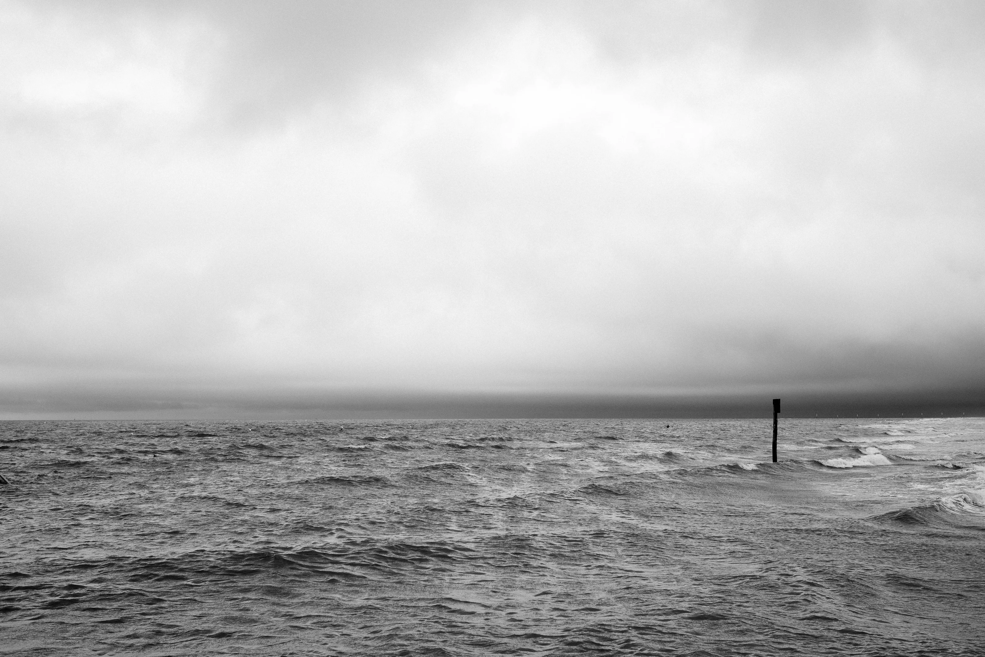 Black and white photo of a vast ocean with a solitary buoy in the distance under a cloudy sky.