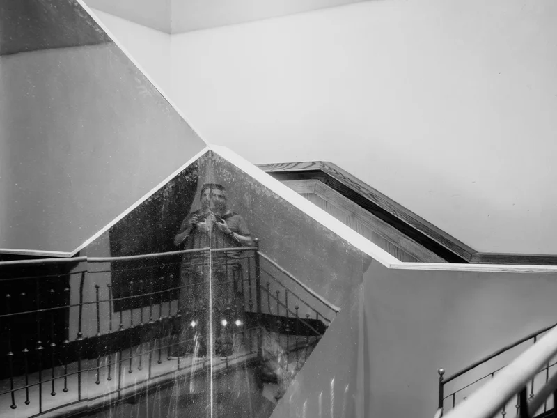 Person taking a photo reflected in a complex, geometric mirror on a staircase.