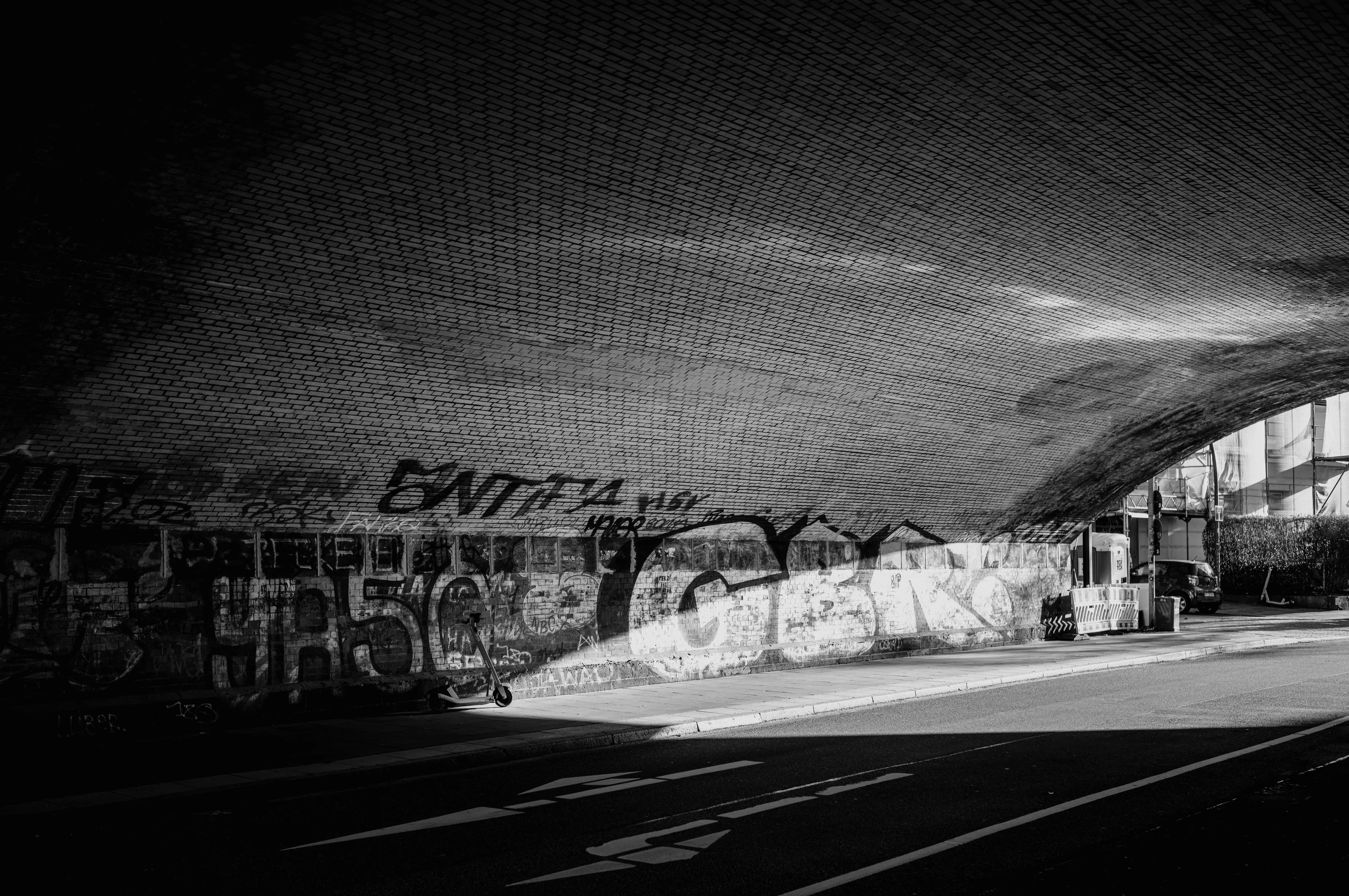Black and white photo of a graffitied brick underpass with sunlight streaming in.