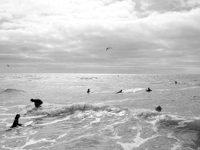 Surfers in the ocean under a cloudy sky with a kite in the background.