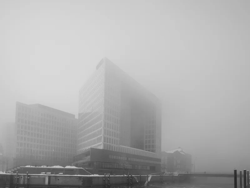 A tall modern building partially obscured by fog looms over a frozen body of water.