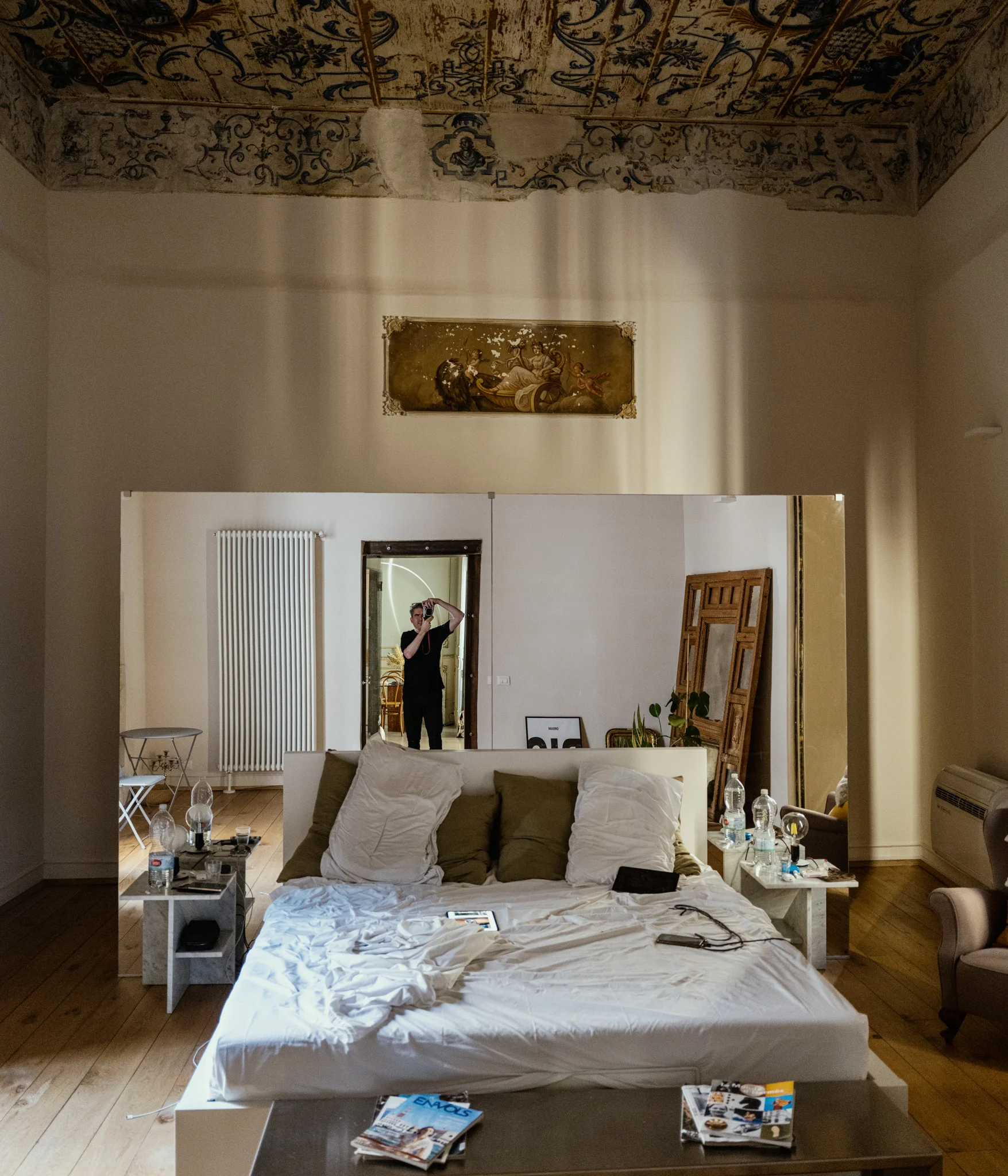 A self-portrait in a mirror inside a stylish bedroom with a large bed and a decorative ceiling, the photographer holding a camera to his eye.
