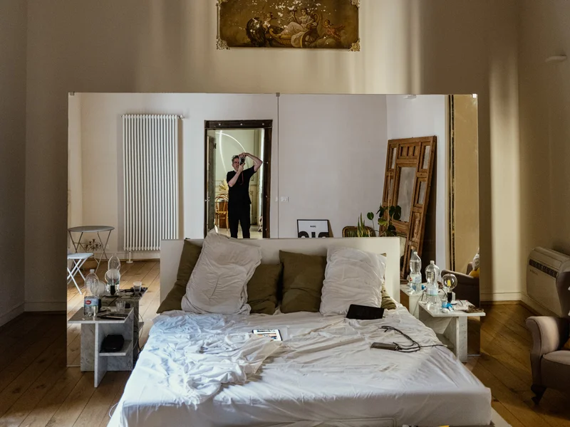 A self-portrait in a mirror inside a stylish bedroom with a large bed and a decorative ceiling, the photographer holding a camera to his eye.