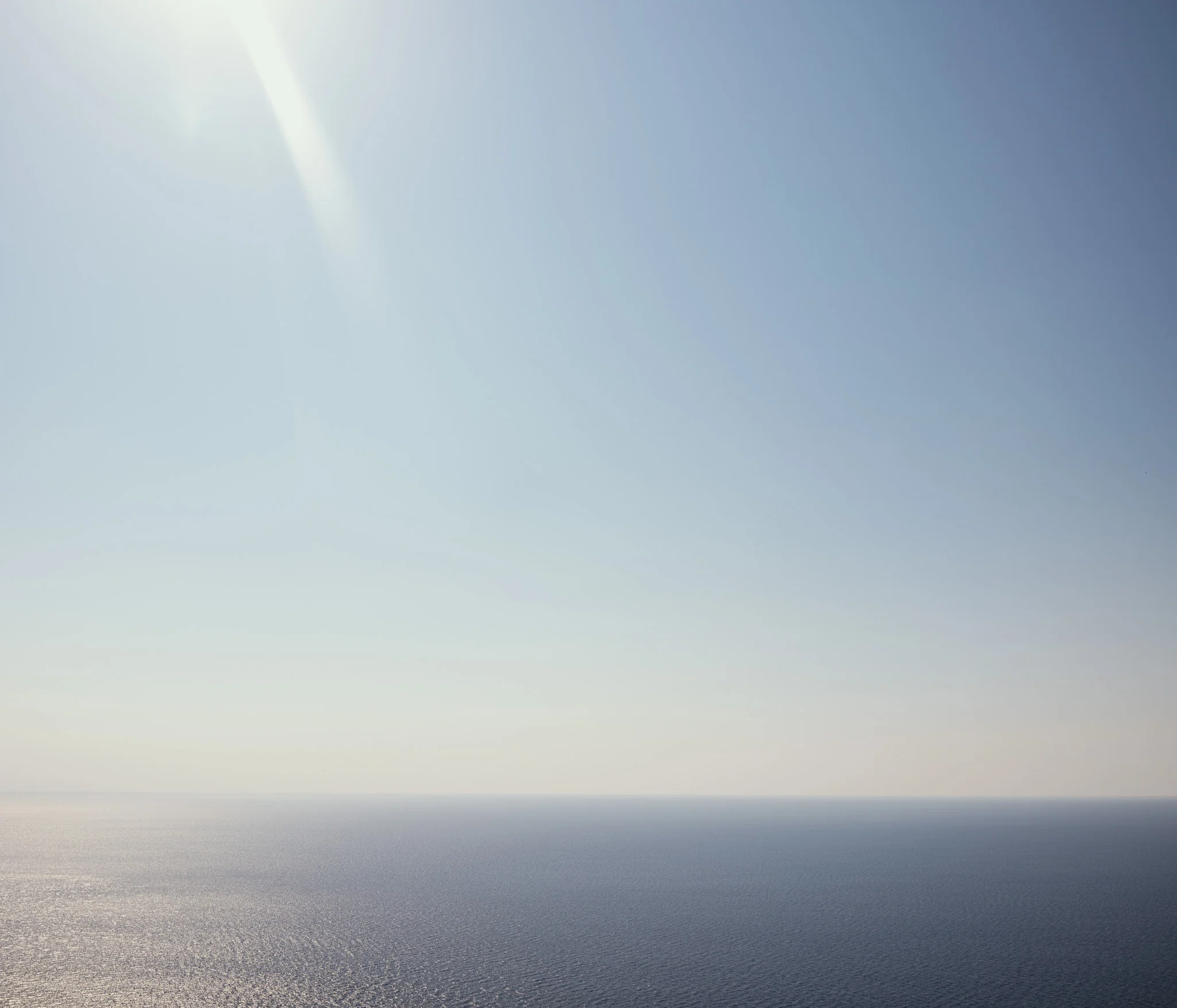 A serene ocean horizon under a clear blue sky with sunlight streaming from the upper left corner.