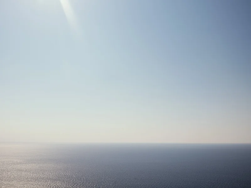 A serene ocean horizon under a clear blue sky with sunlight streaming from the upper left corner.