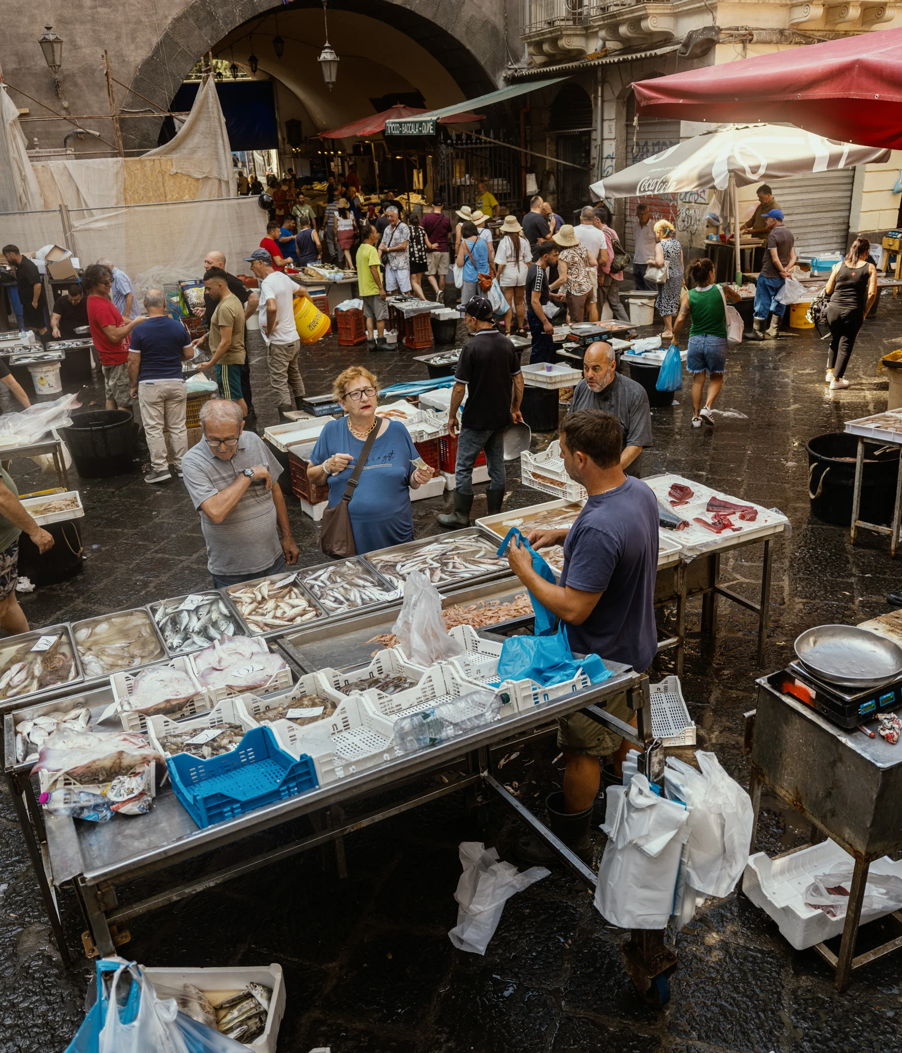 Bustling outdoor fish market with people shopping and vendors displaying seafood.