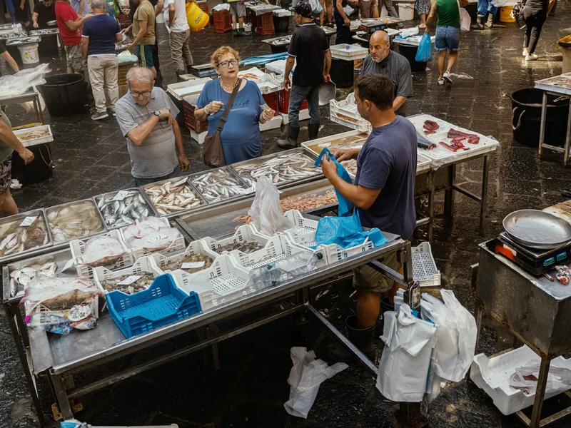 Bustling outdoor fish market with people shopping and vendors displaying seafood.
