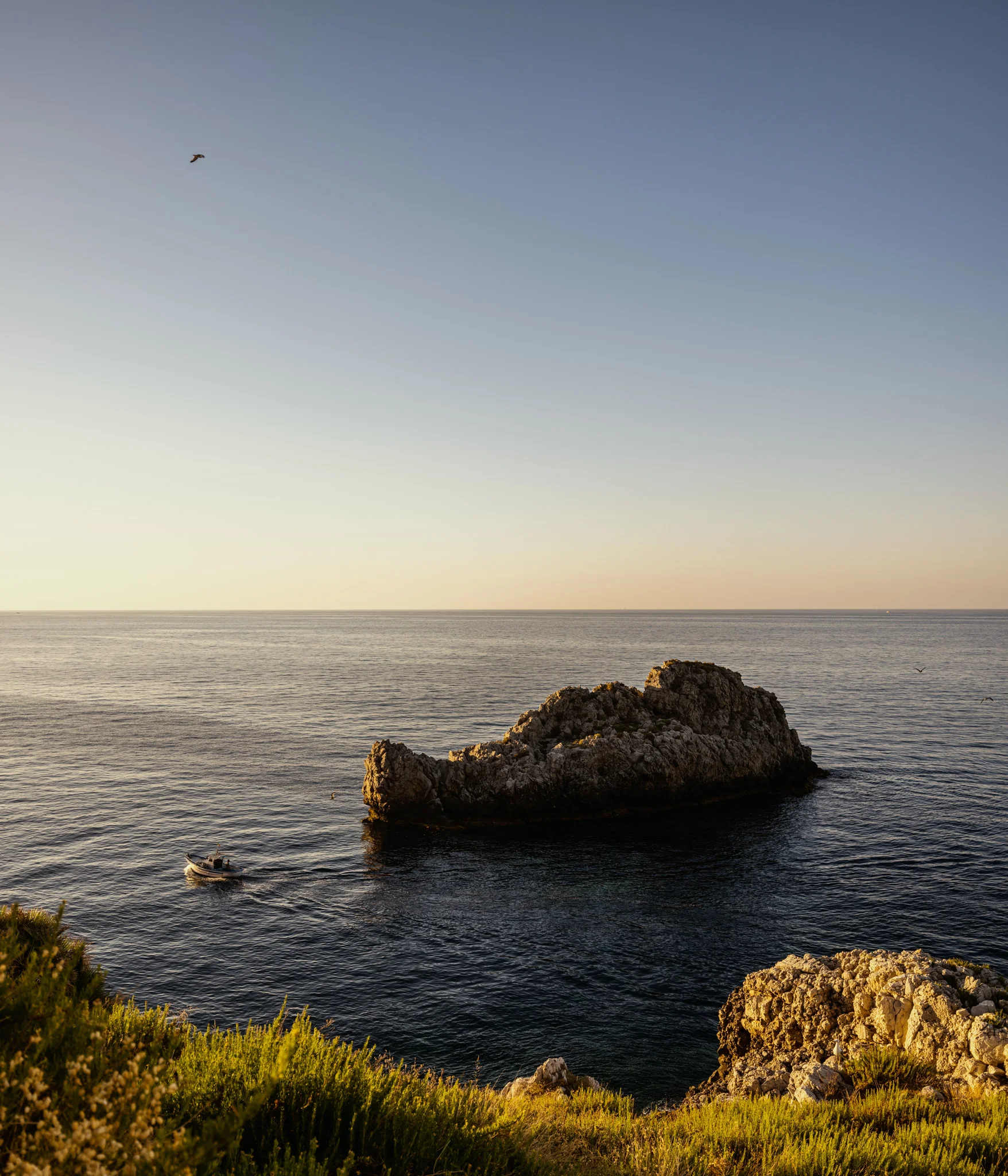 A boat navigates calm waters near a rocky outcrop under a clear sky.