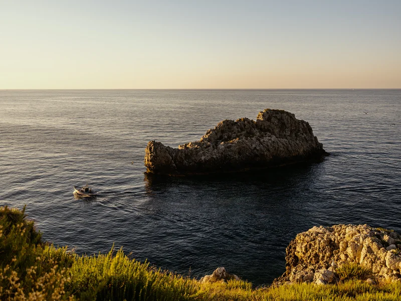 A boat navigates calm waters near a rocky outcrop under a clear sky.