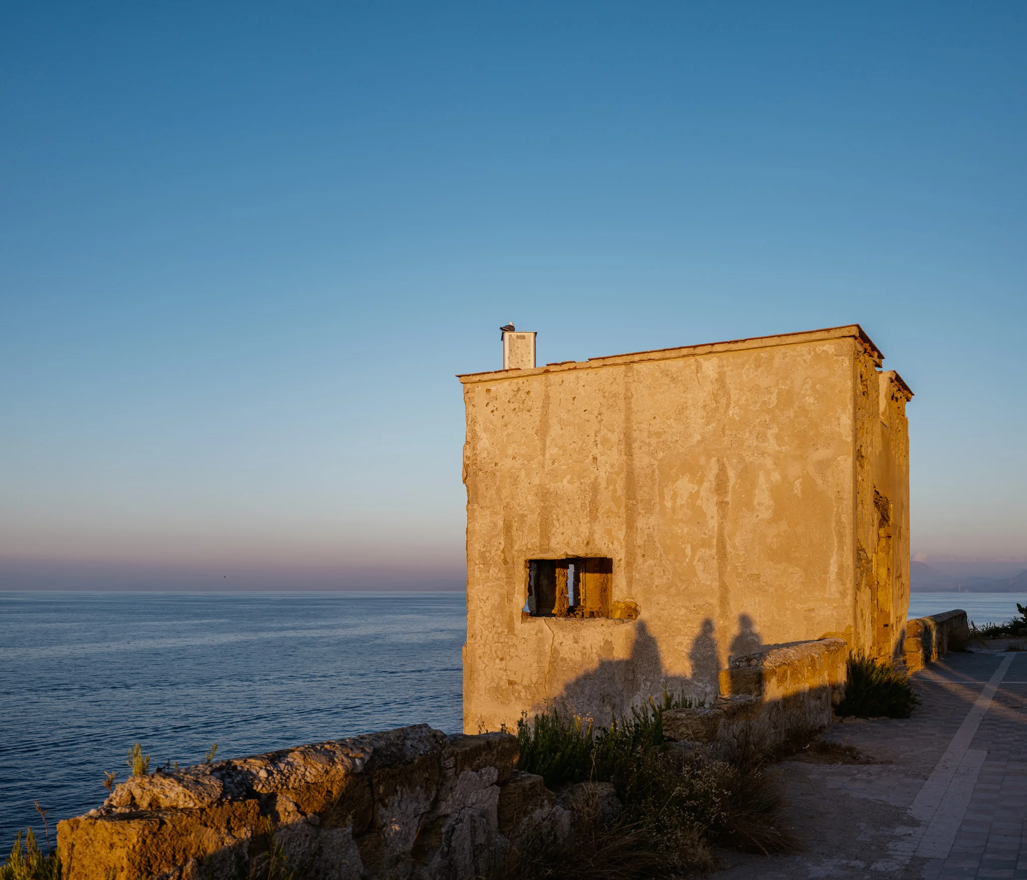 A weathered seaside building with evening light casting long shadows on its wall.