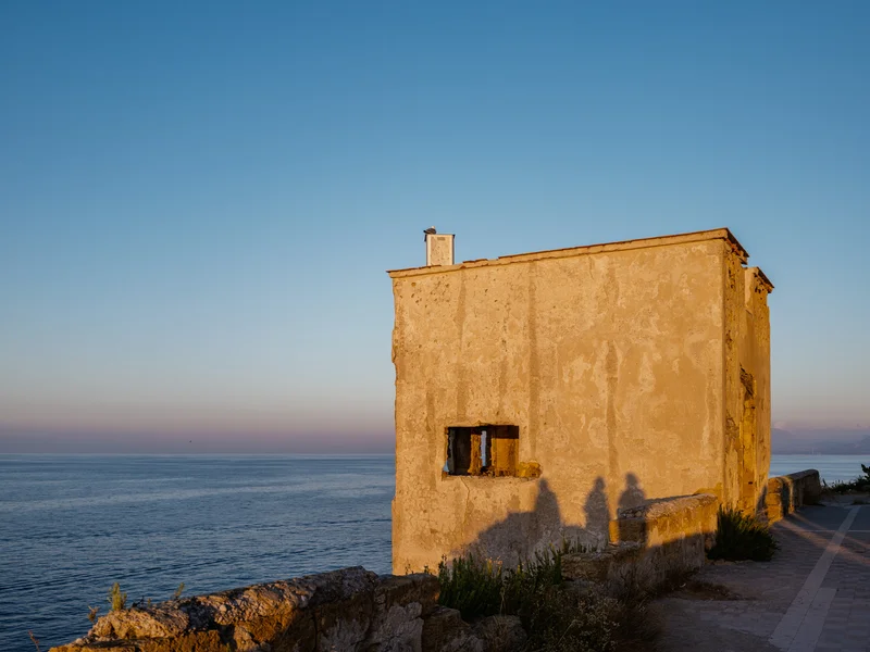 A weathered seaside building with evening light casting long shadows on its wall.
