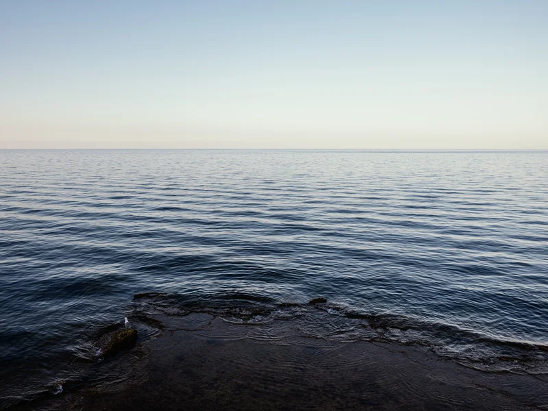 A calm ocean under a clear sky with a rocky shore in the foreground.