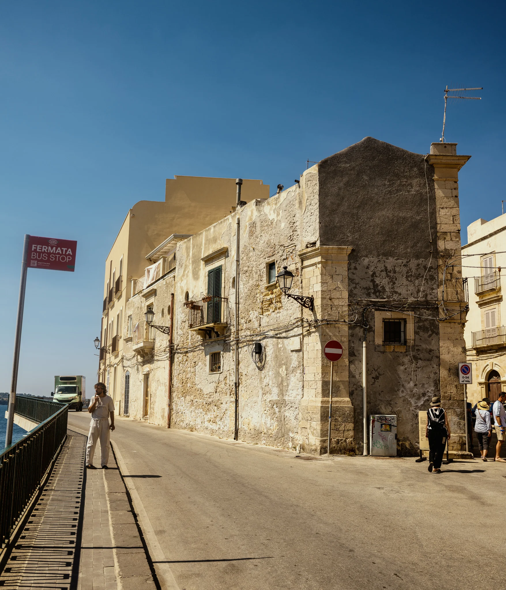 Street scene with aged buildings in sunlight, featuring a bus stop sign and pedestrians.