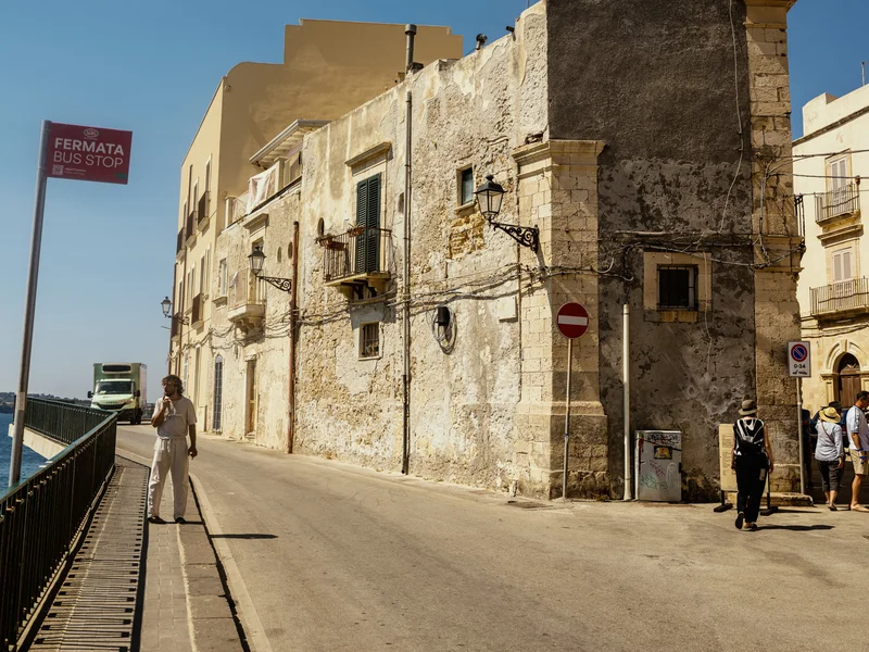 Street scene with aged buildings in sunlight, featuring a bus stop sign and pedestrians.