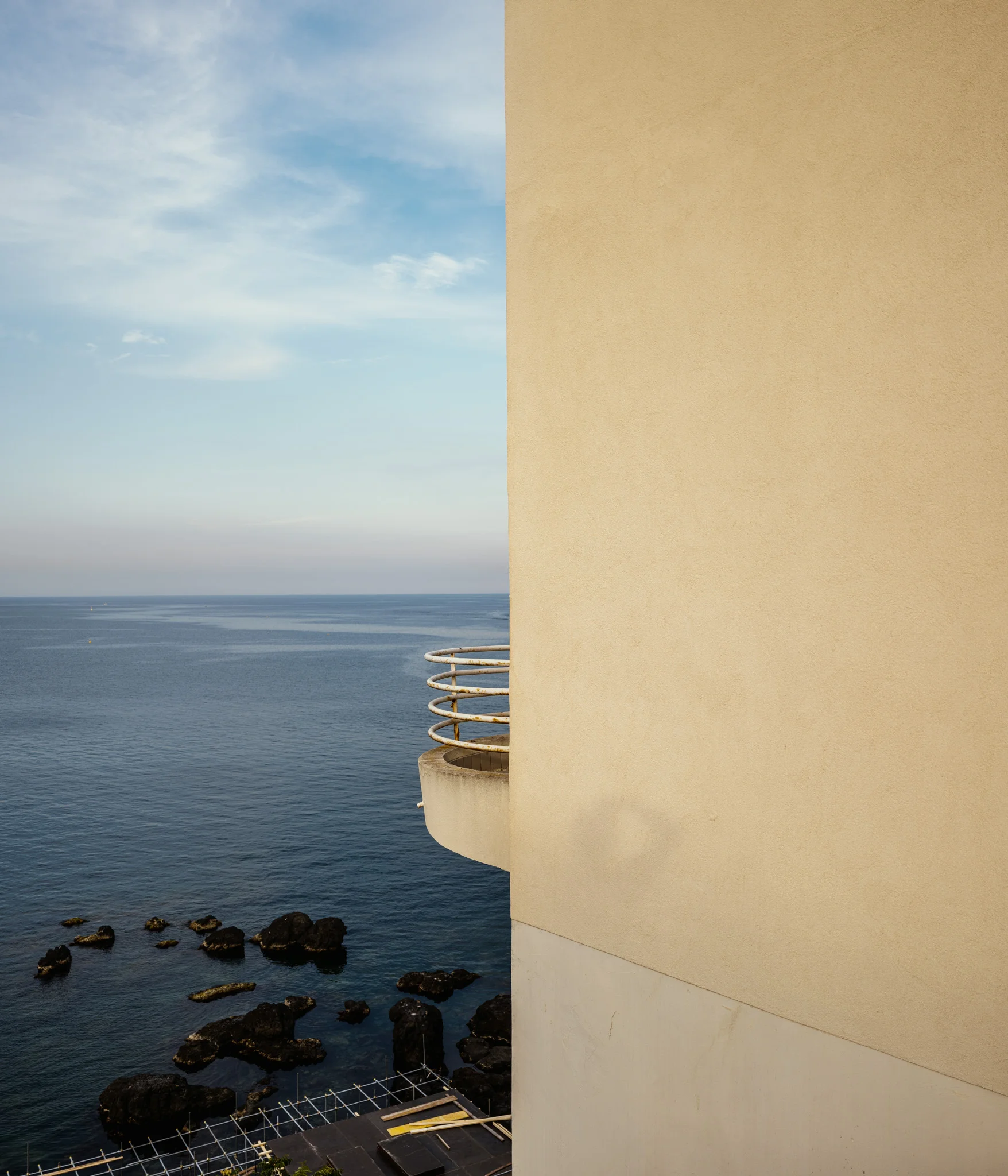 A beige building with a curved balcony overlooks a calm sea with scattered rocks.