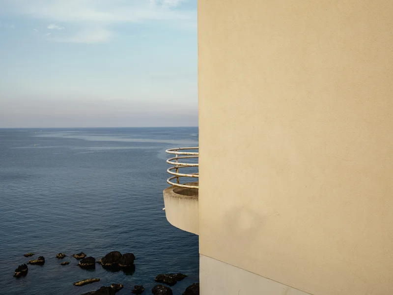 A beige building with a curved balcony overlooks a calm sea with scattered rocks.