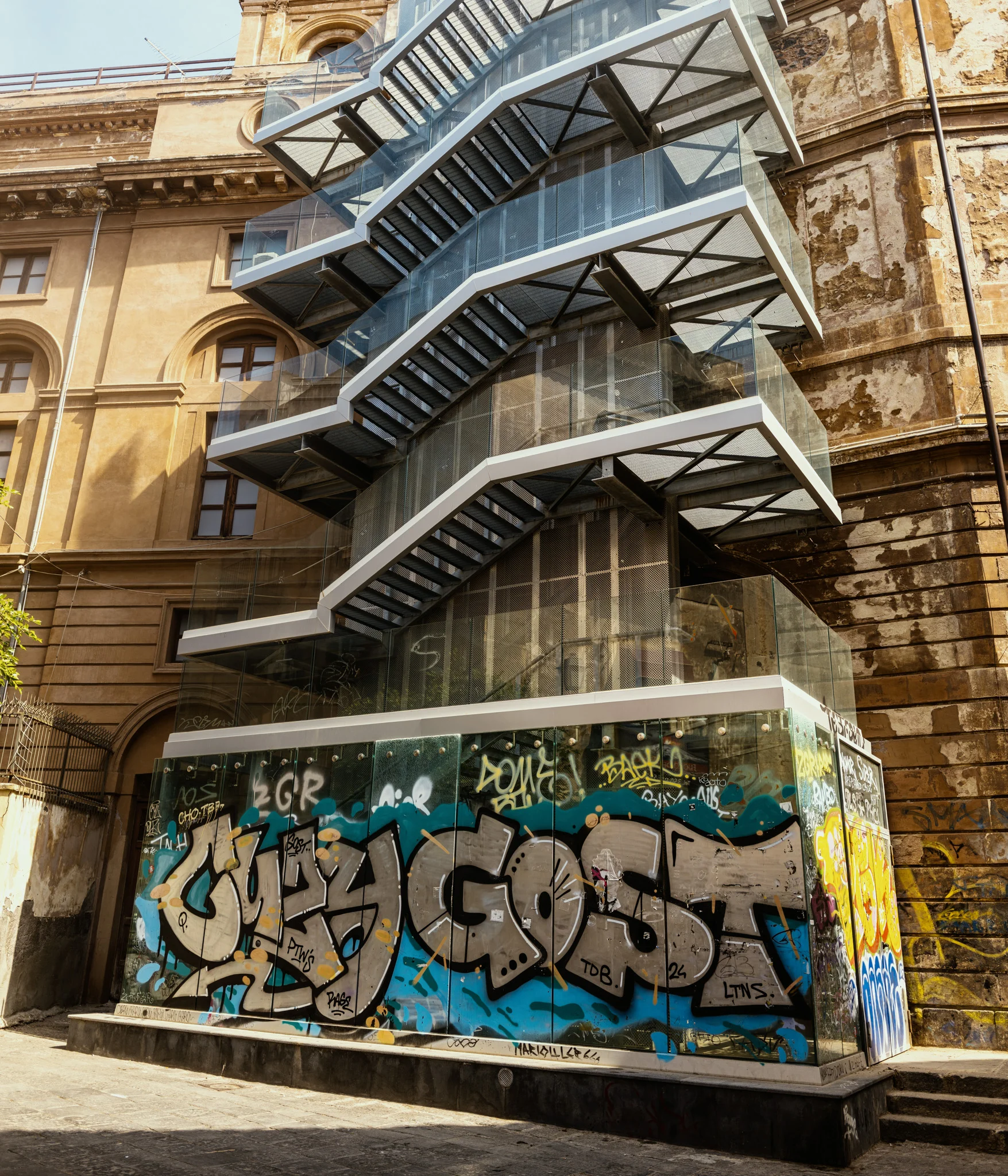 Multi-level glass staircase with graffiti-covered wall in an urban setting.