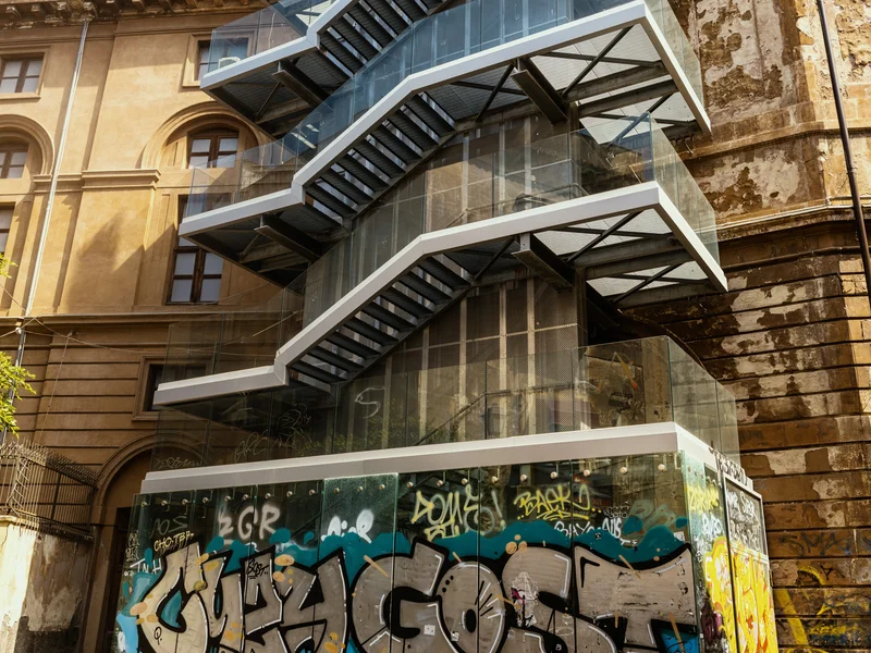 Multi-level glass staircase with graffiti-covered wall in an urban setting.